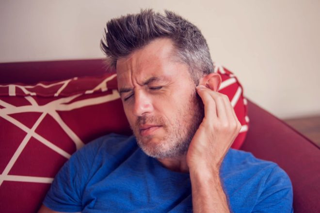 A young man with pain on his face puts a hand on his ear to try and relieve his tinnitus pain symptoms.